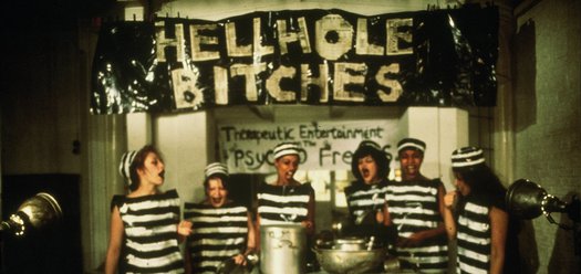 6 prisoners standing in front of cooking pots with a banner behind them reading 'Hellhole Bitches' - still from Scrubbers (1982)
