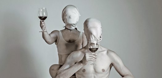 Two scantily clad figures - a male and female - in white clothing, white body paint , and white masks covering their entire heads. They are both holding wine glasses. The female figure is raising her glass and the male figure is sniffing the wine.