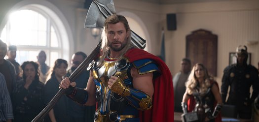 Chris Hemsworth as Thor, standing in a room surrounded by people. He's wearing a gold and blue suit of armour and a flowing red cape. He's holding the axe stormbreaker, made from tree branch and a jagged blade. His hair is long and framing his face.