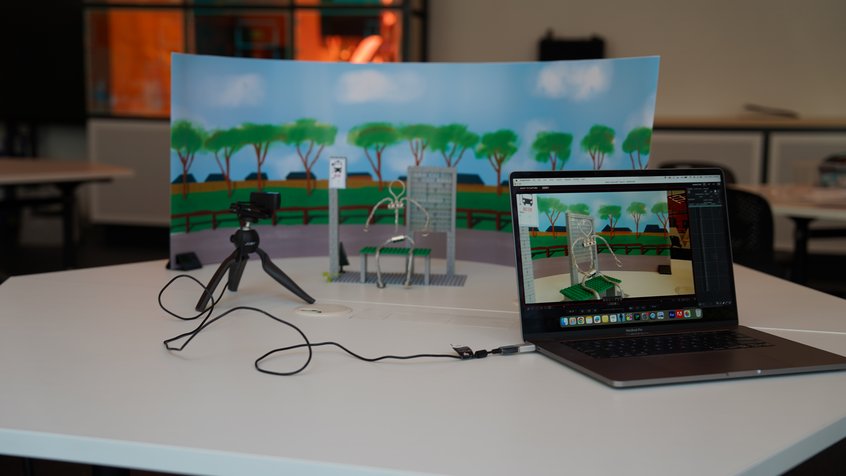 A stop motion animation setup, featuring a laptop, webcam on a small tripod, as well as an animation set with an armature character, bus stop, and pre printed background