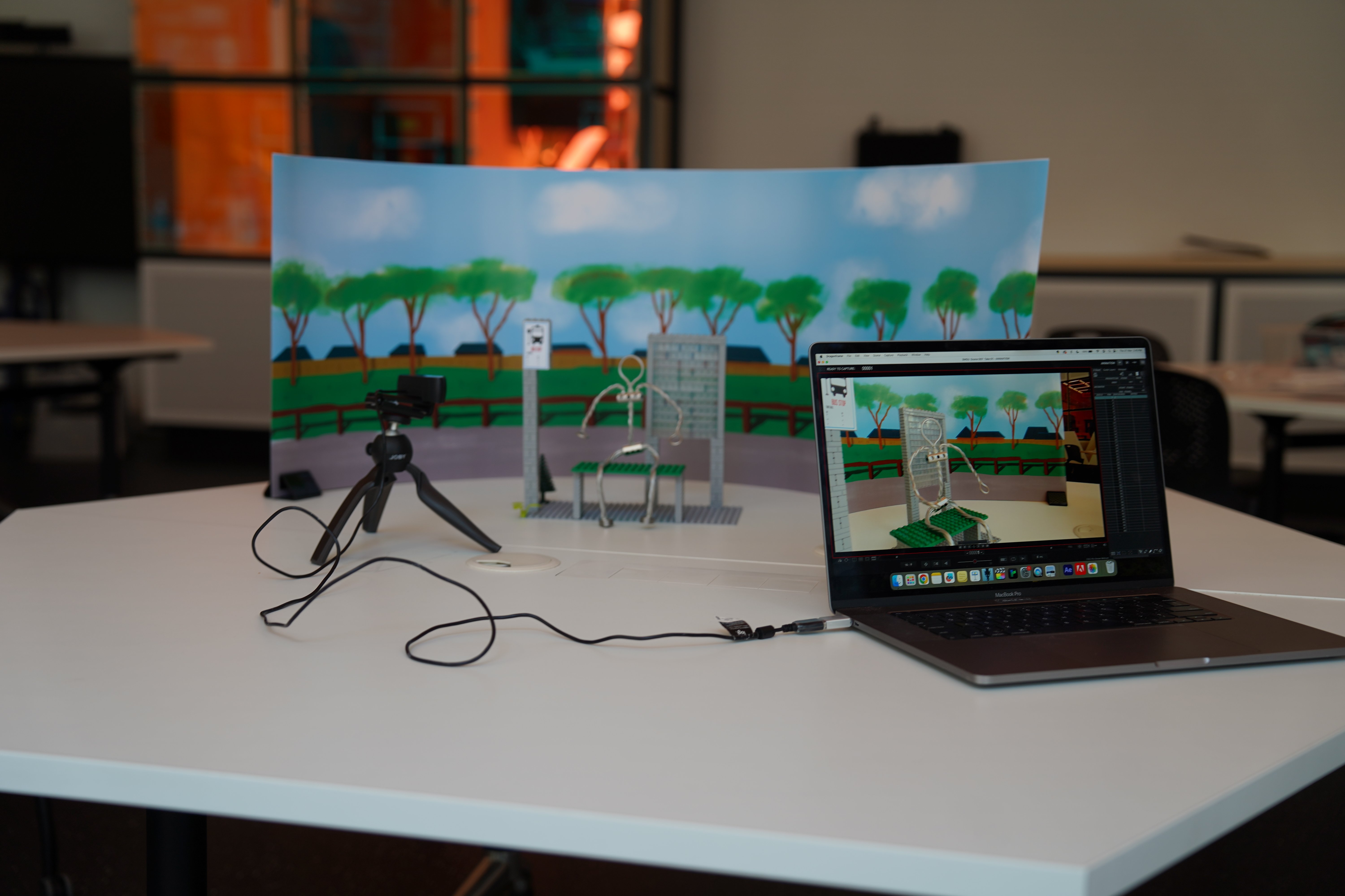 A stop motion animation setup, featuring a laptop, webcam on a small tripod, as well as an animation set with an armature character, bus stop, and pre printed background
