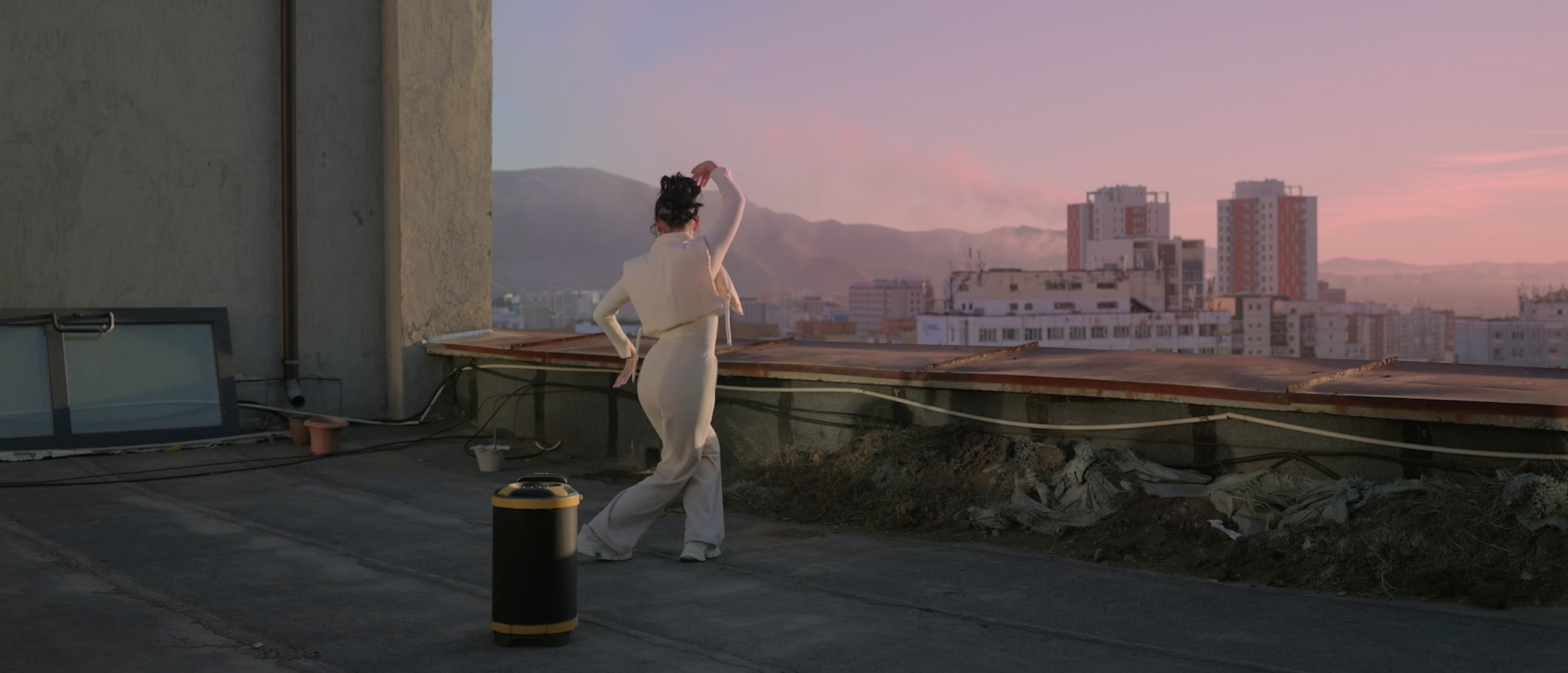 Young person dances on roof looking out onto pink sky, city and mountains