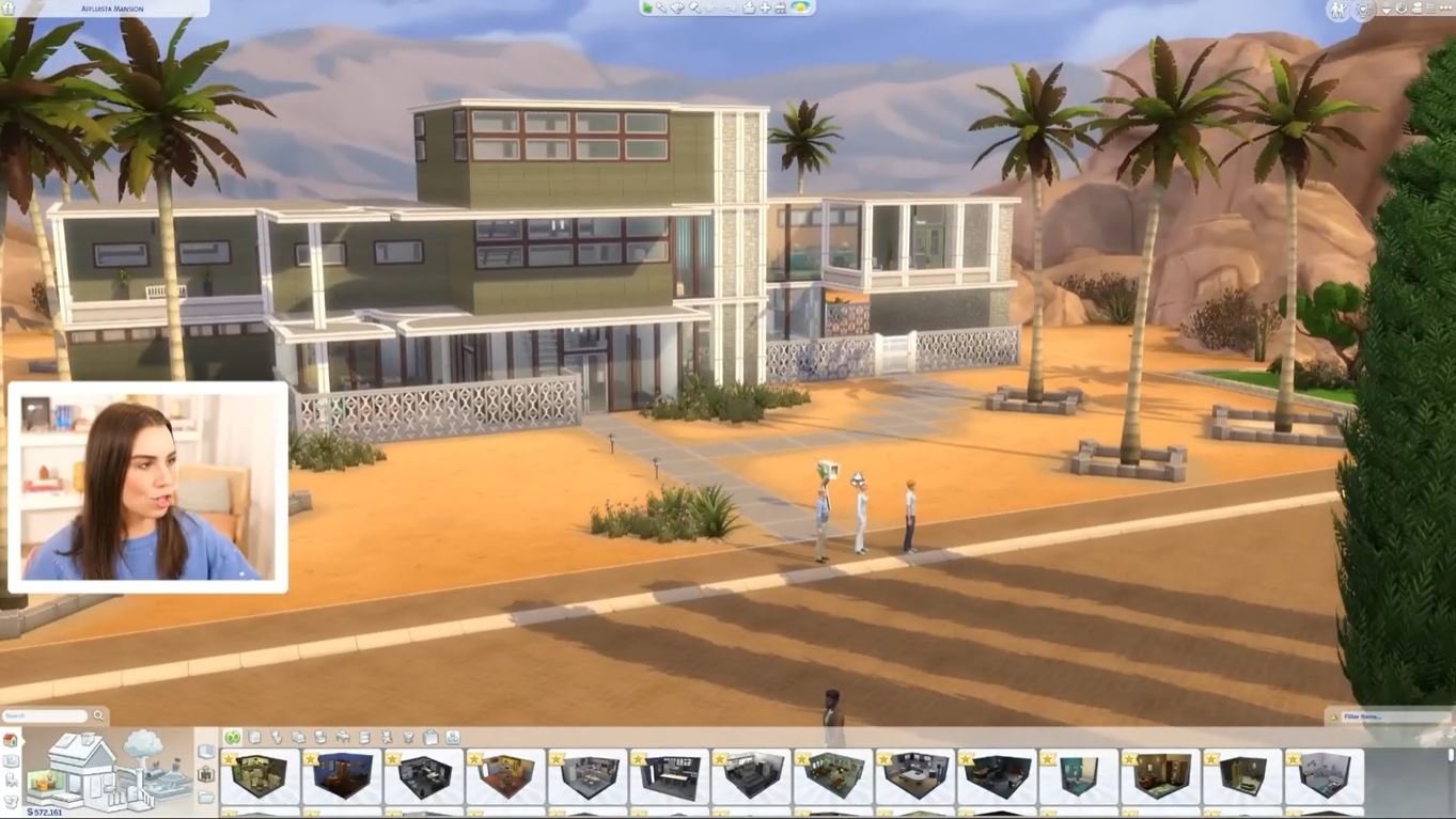 A livestream of a sims build video, showing the simmer deligracy, a brunette woman in a blue sweater, explaining the large white mansion that shes building.