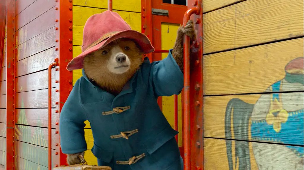 Large animated bear character wearing a hat and a coat hangs onto handle of train door
