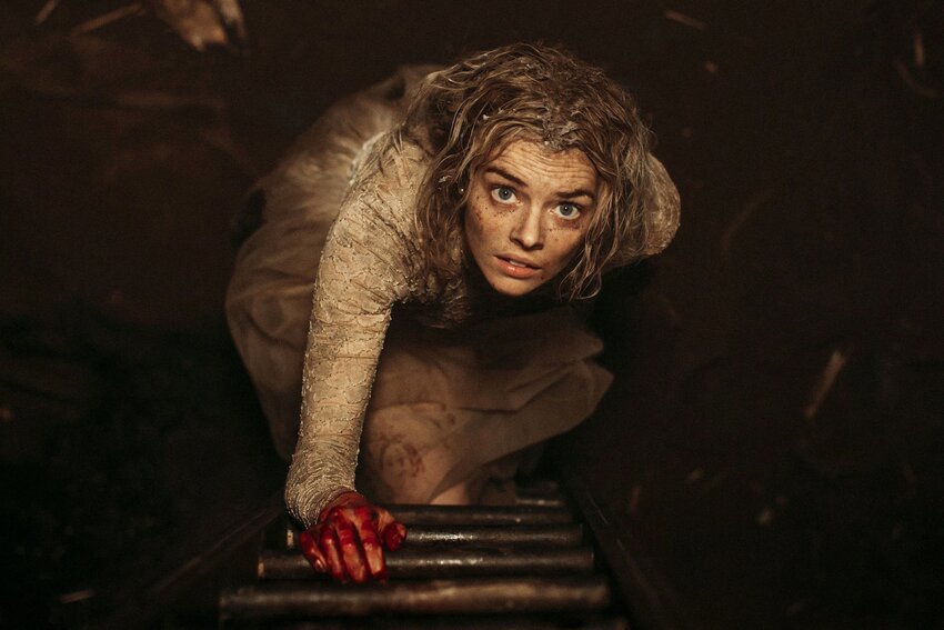 A scared woman in a bridal dress, covered in mud and blood, looking up at the camera, as if about to climb a ladder. Stringy blonde hair frames her face and her eyes are wide with shock but a steely determination to survive.
