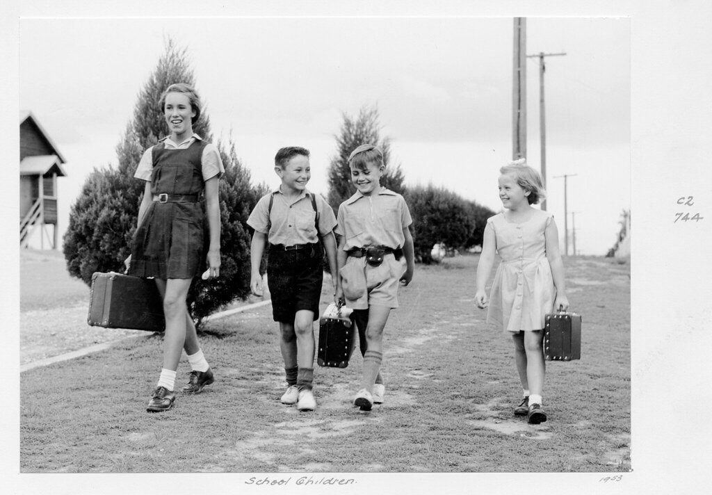 Four smiling children with schoolbags walk along country road in 1950s. The image is black and white.