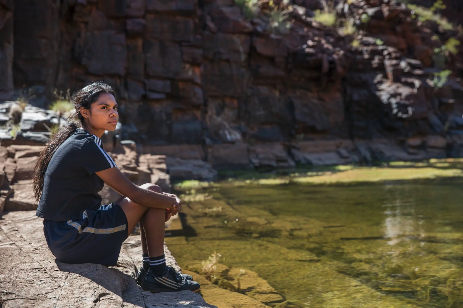 The character of Murra from Sweet As, dressed casually, sits by a water hole in a beautiful, Western Australian landscape.