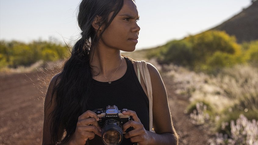 A young Indigenous Australian girl (Shantae Barnes-Cowan) holding a camera and looking to her right in a still from the film Sweet as (2022).