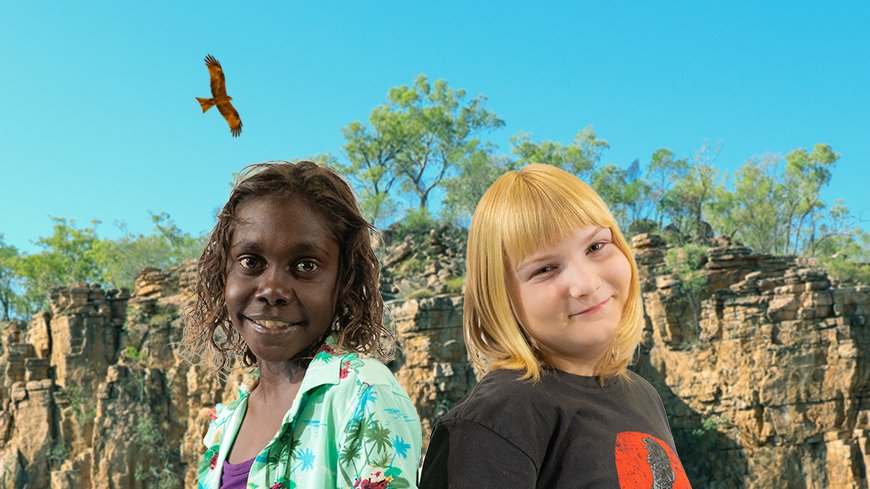 Two smiling children stand against background of rocks, trees and sky with an eagle flying above them.