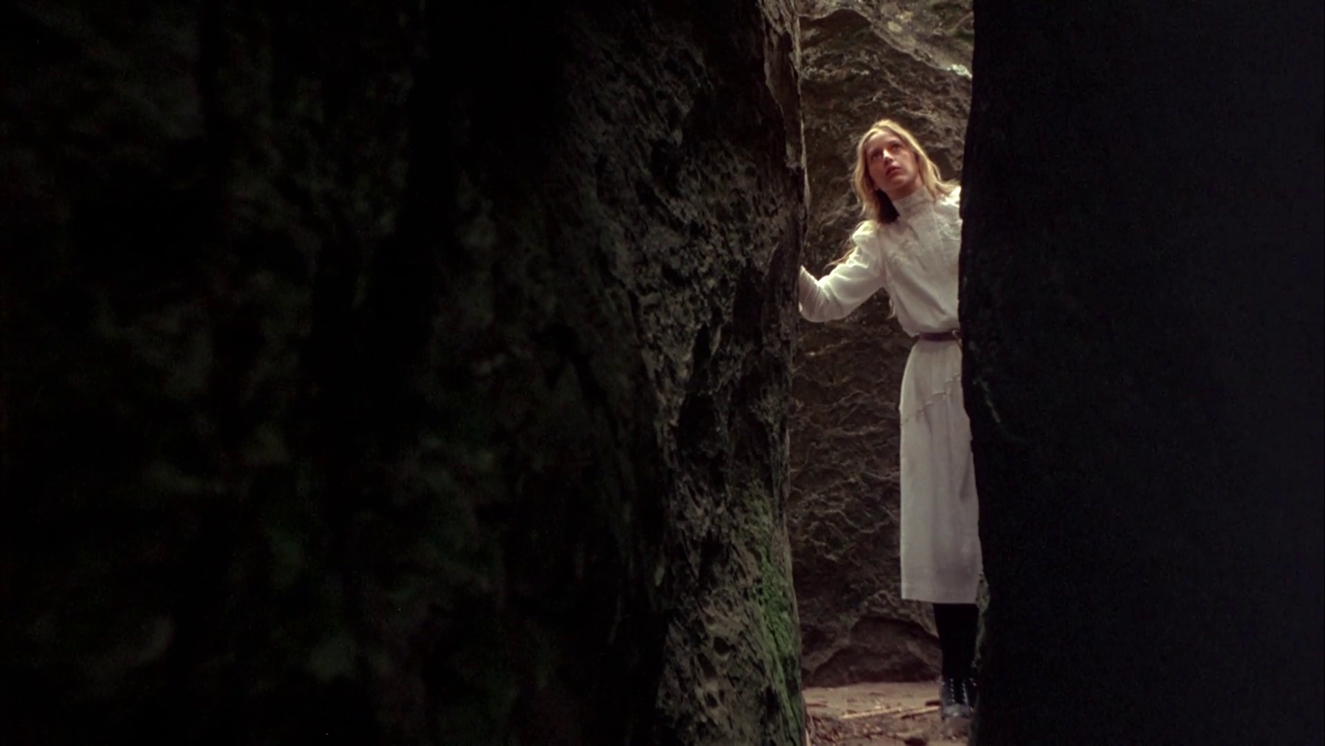 The character of Miranda from Picnic at Hanging Rock stands between two large boulders looking up.