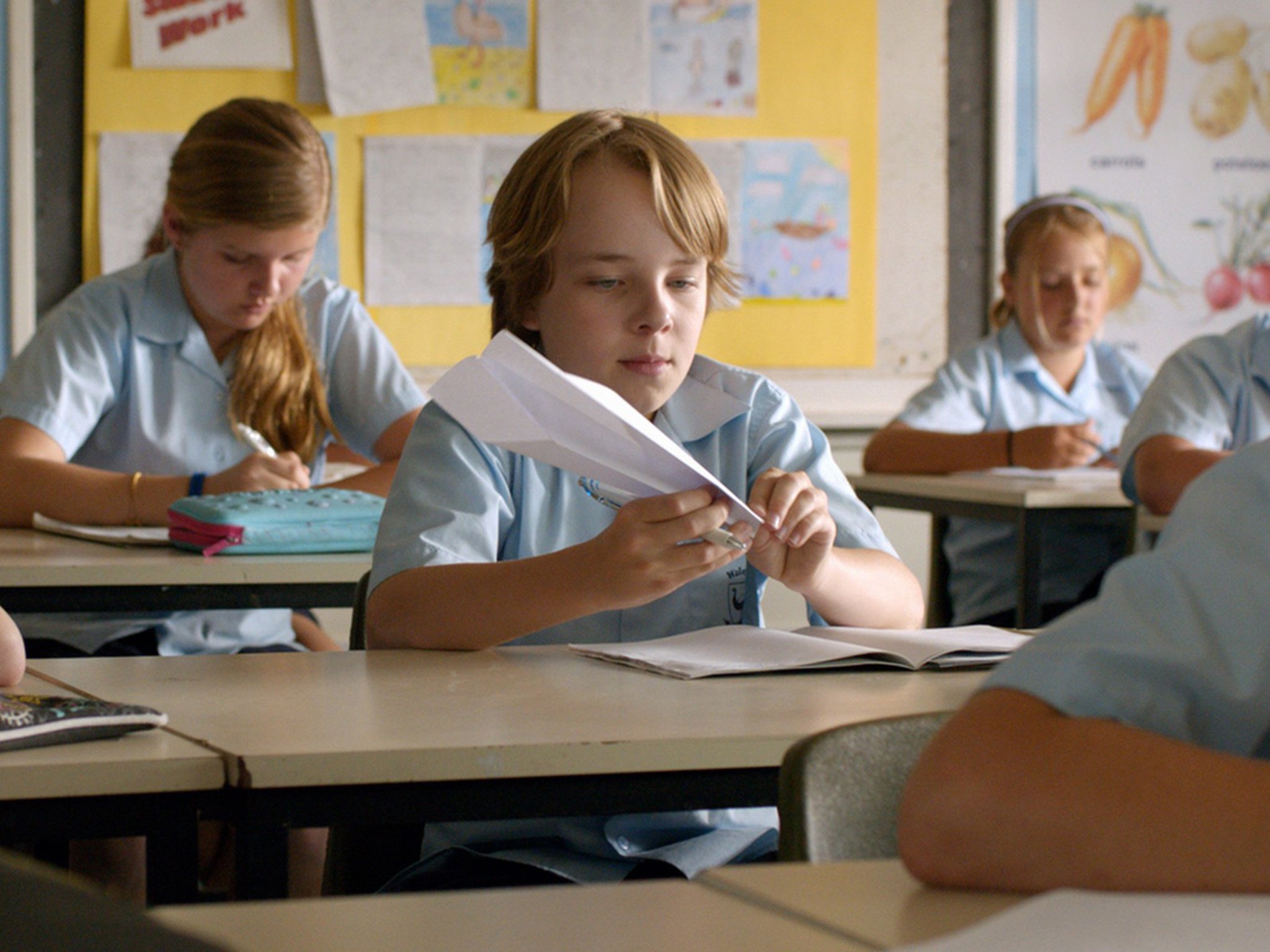 School boy sits at desk in a classroom making a paper plane