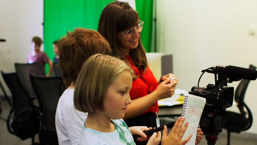 Kids holding a script and directing a film behind the camera with an instructor looking on, at ACMI