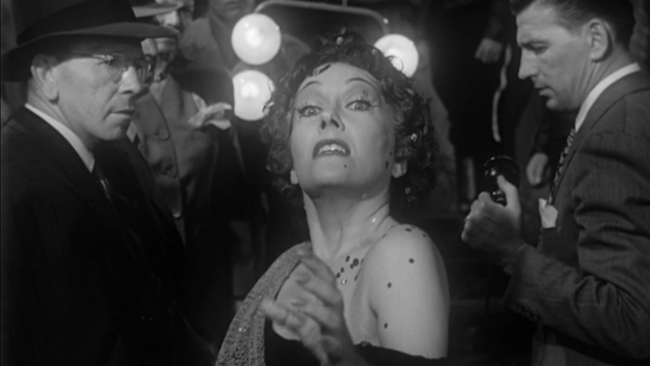 An image of a woman in stark white make-up and a regal hollywood hairstyle, recoiling from the camera but edging towards it, eyes wide, hands outstretched. She wears a formal gown. Behind her are two men and a film crew with lights.
