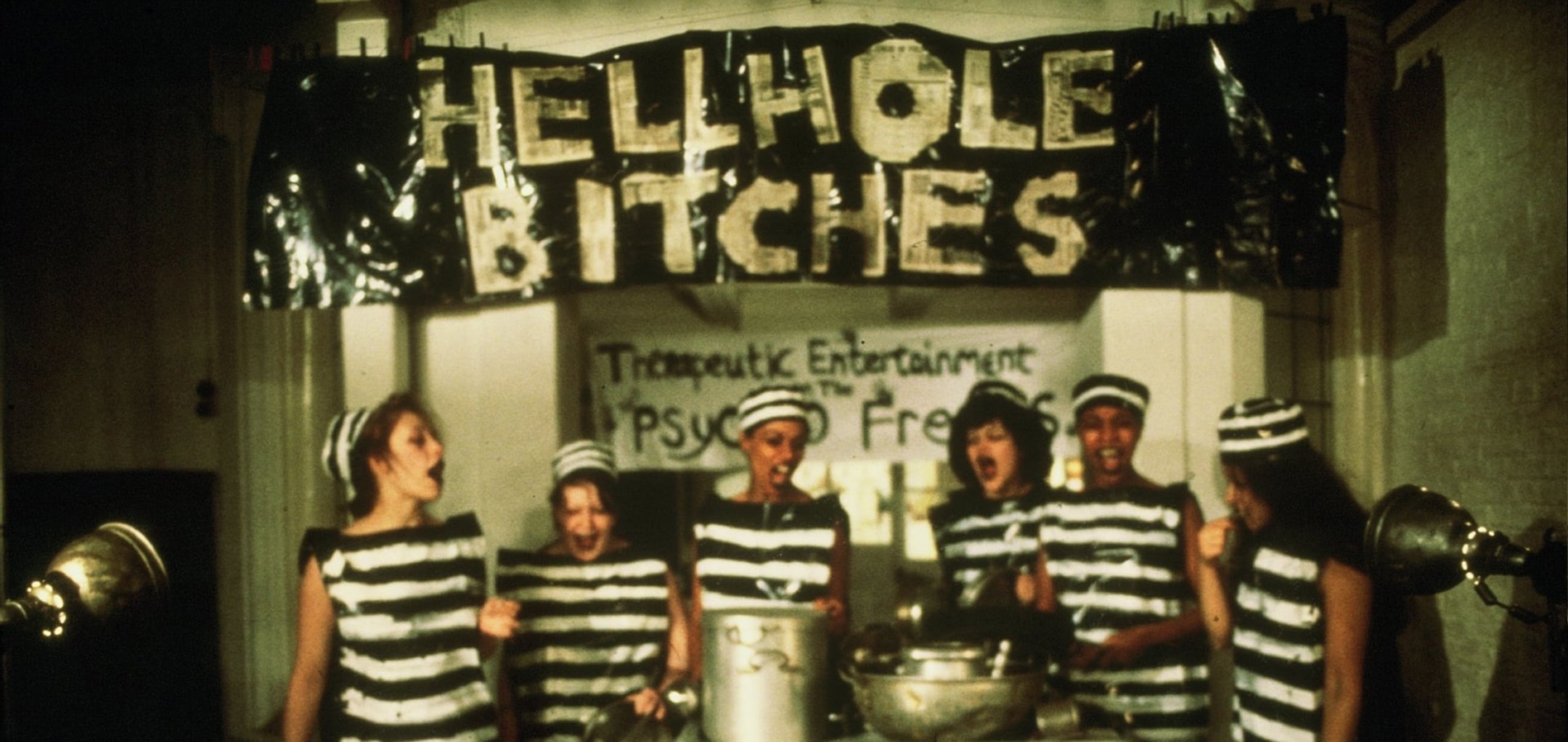 6 prisoners standing in front of cooking pots with a banner behind them reading 'Hellhole Bitches' - still from Scrubbers (1982)