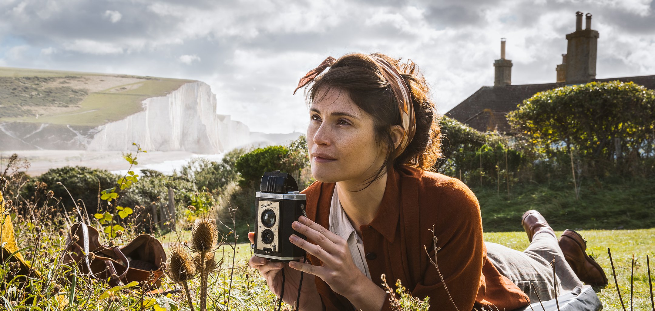 Gemma Arterton lying in a field with a camera in a still from Summerland (2020)