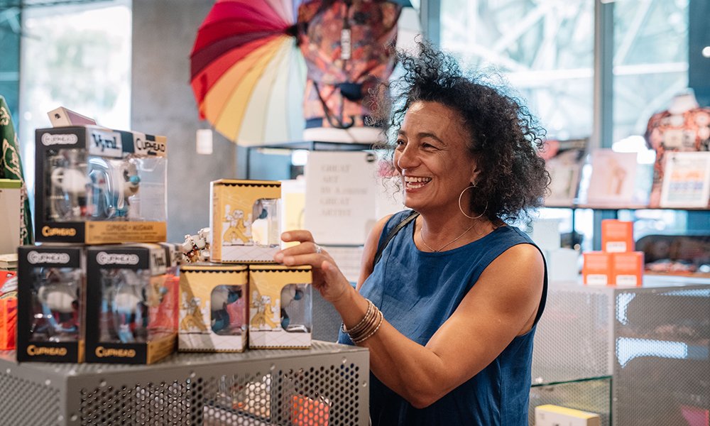 A woman browses items in the ACMI Shop. She is wearing a blue top and smiling. Photograph by Phoebe Powell