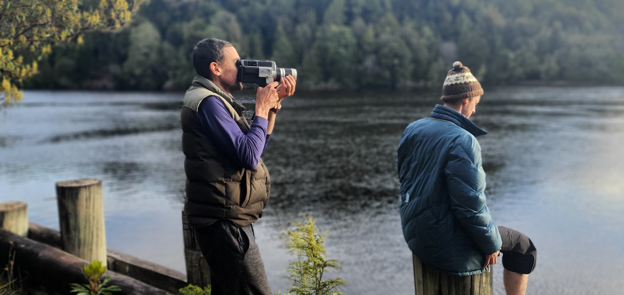 Still from the 2021 documentary 'Battle on the Franklin'. Two men stand on a picturesque riverbank overlooking the Franklin River in Tasmania. Both are wearing cold weather jackets and the one on the left is holding a video camera to his eye.