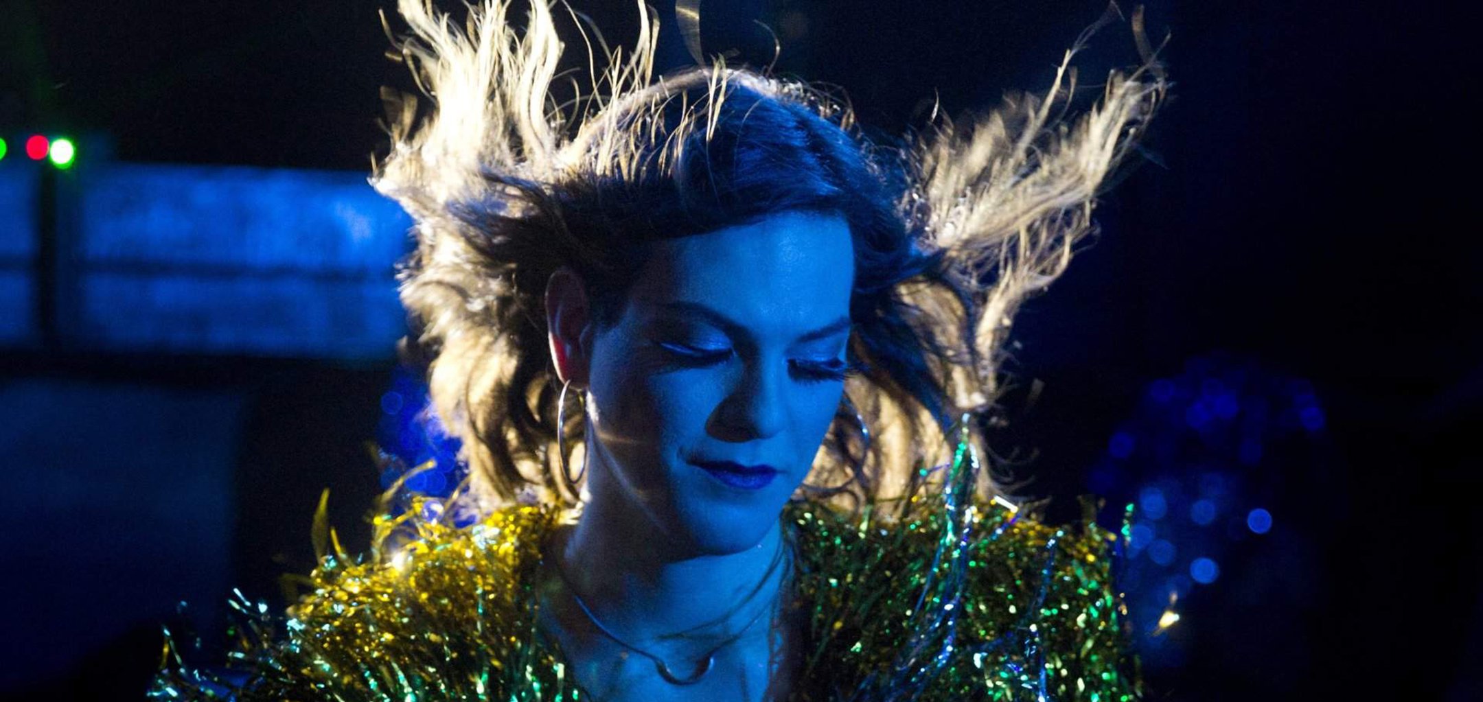 A medium shot of Daniela Vega in the film 'A Fantastic Woman' (2017). The photo was taken at night and her hair is blown upwards by the wind and her face tinted blue in the light. She wears a green and gold, shiny shawl around her shoulders.
