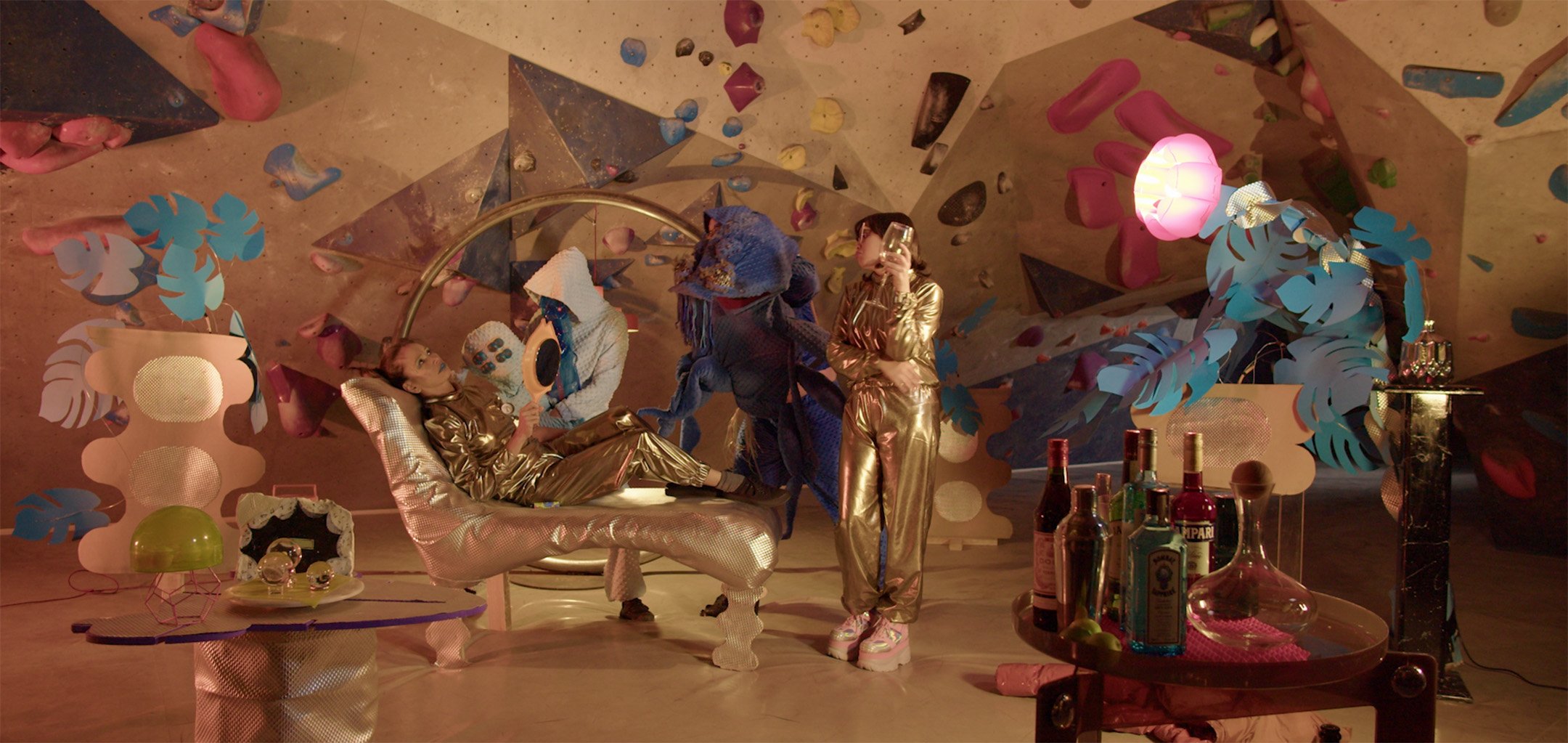 A mainly golden stage set with angular walls and colourful decorations. Two women in gold suits stand in the middle - one is reclining on a gold chair and the other is standing and holding a wine glass.