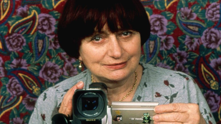 Agnès Varda resting a camcorder on top of a painting with a floral background
