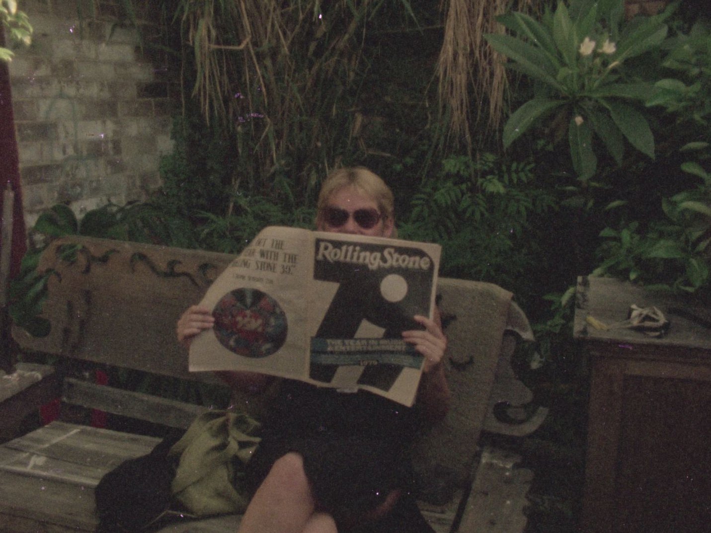 Sue Ford sitting in a garden, wearing sunglasses and peering out behind a copy of Rolling Stone she is holding.