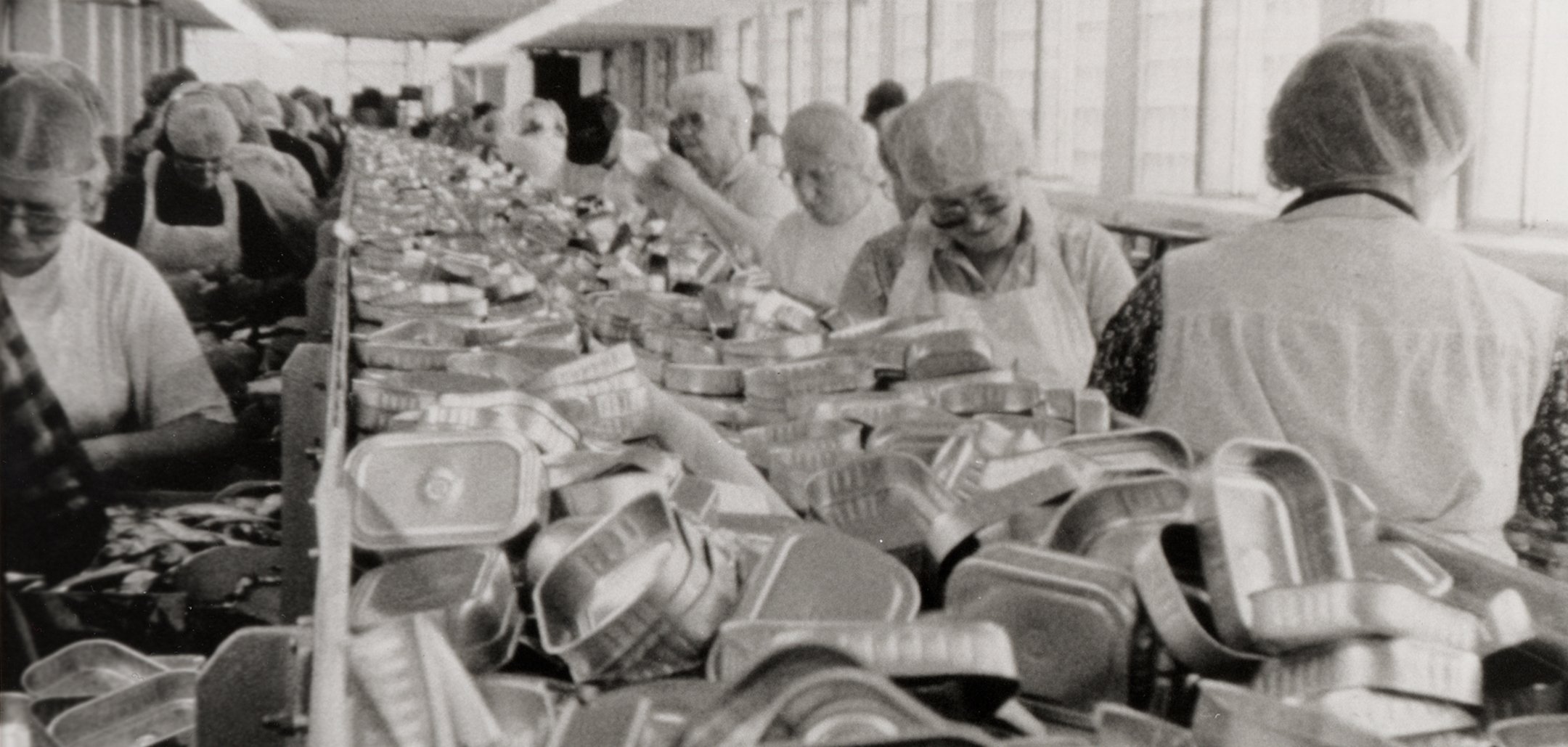 A black and white photo of empty sardine cans going down a conveyor belt, either side of which stand female factory workers. Still from 'Belfast, Maine' (1999)