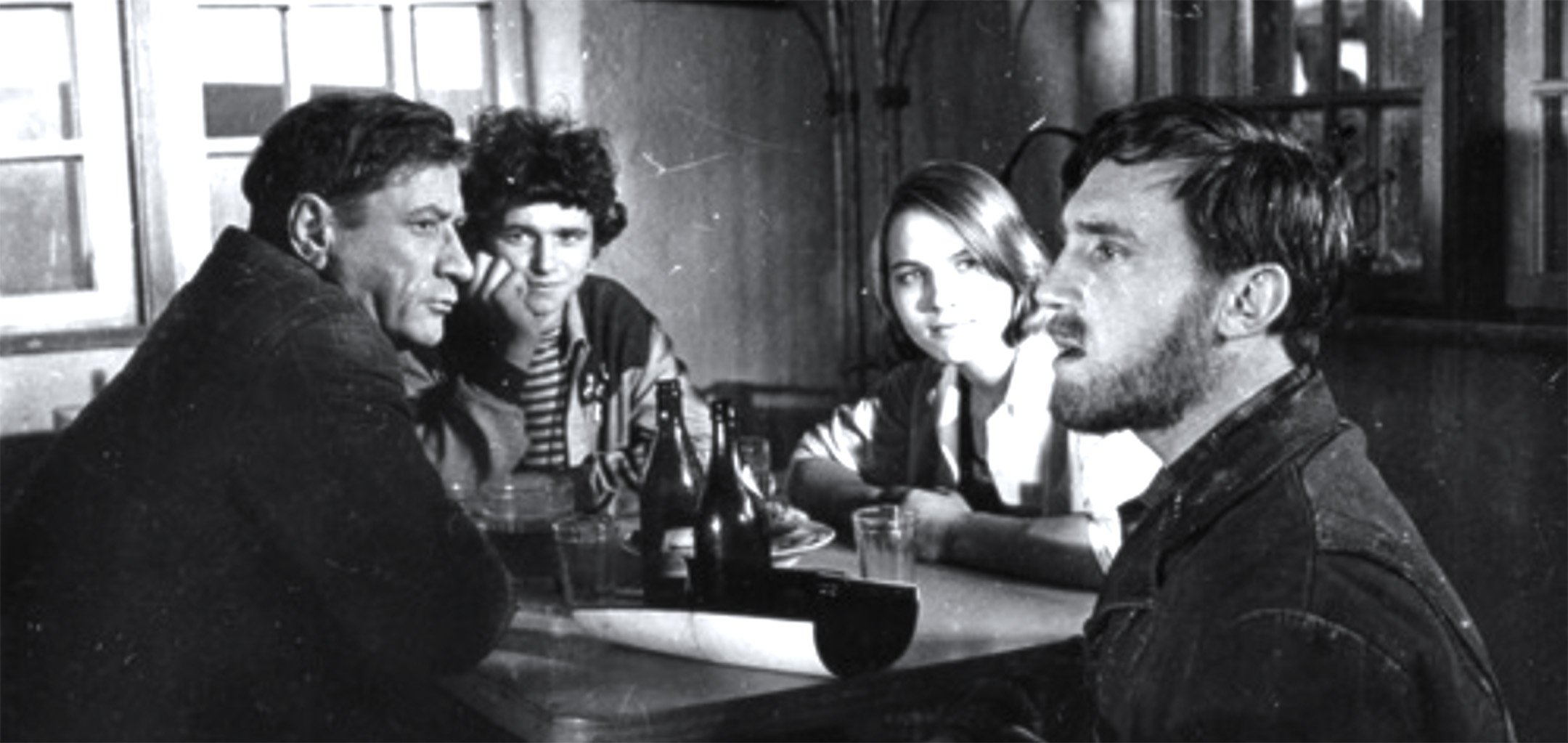 A black and white still of a group of Ukrainian people sitting around a table. The man in the foreground is looking off into the distance and singing.