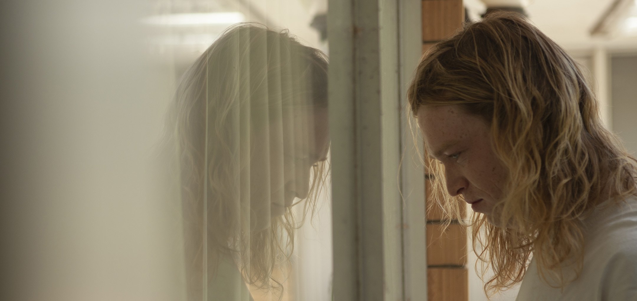 A side view of a young man (Caleb Landry Jones as Nitram) with long blonde hair looking downwards into a reflective window. A still from 'Nitram' (2021) directed by Justin Kurzel.