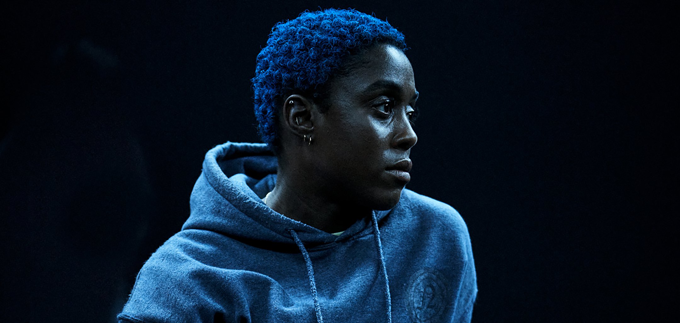 A close, side profile shot of Lashana Lynch in the film 'ear for eye' (2021) directed by debbie tucker green. Lynch is wearing a grey hoodie and has short, curly hair dyed blue.