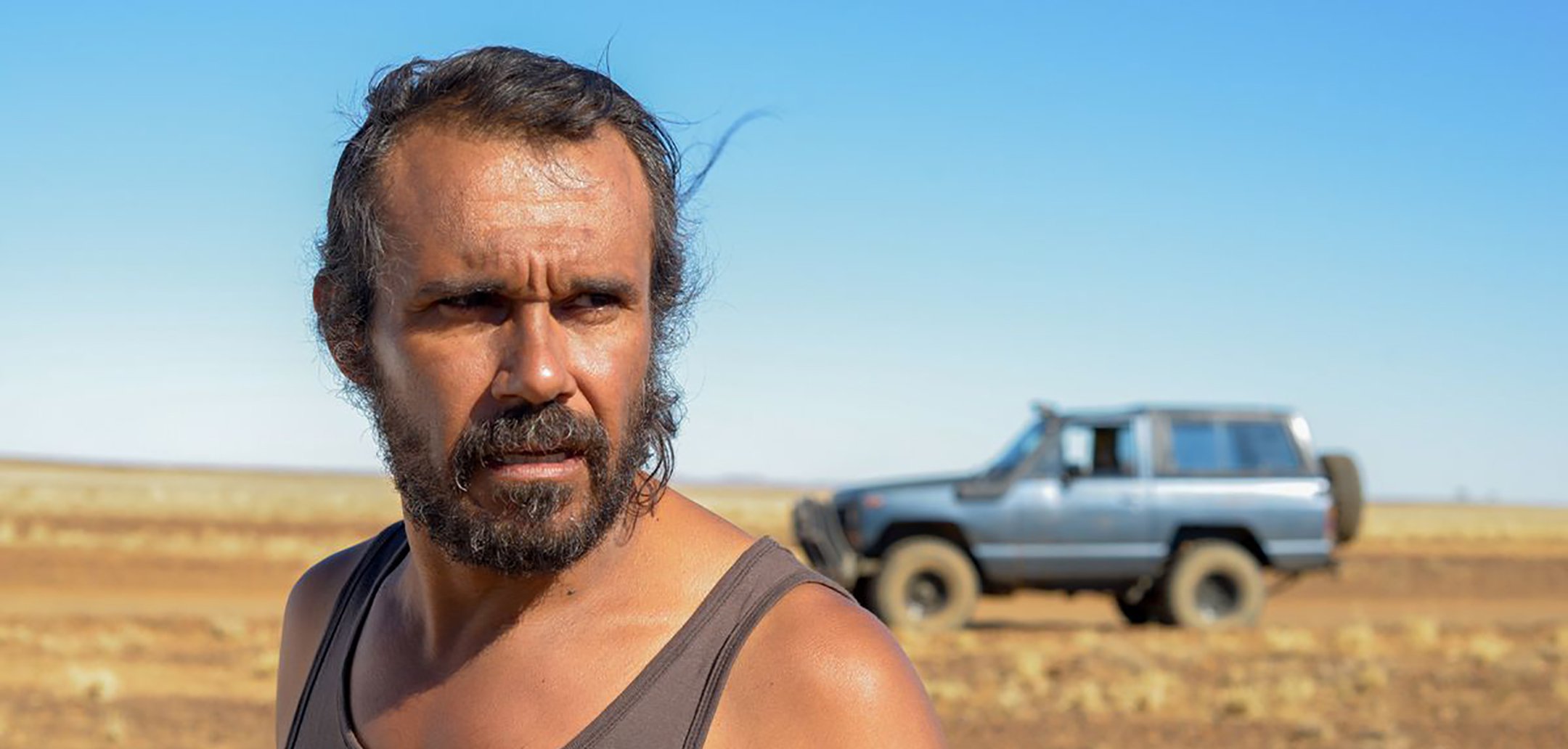 Closeup of Aaron Pedersen in the desert, in the background there is a jeep. Still from 'Goldstone' (2016)