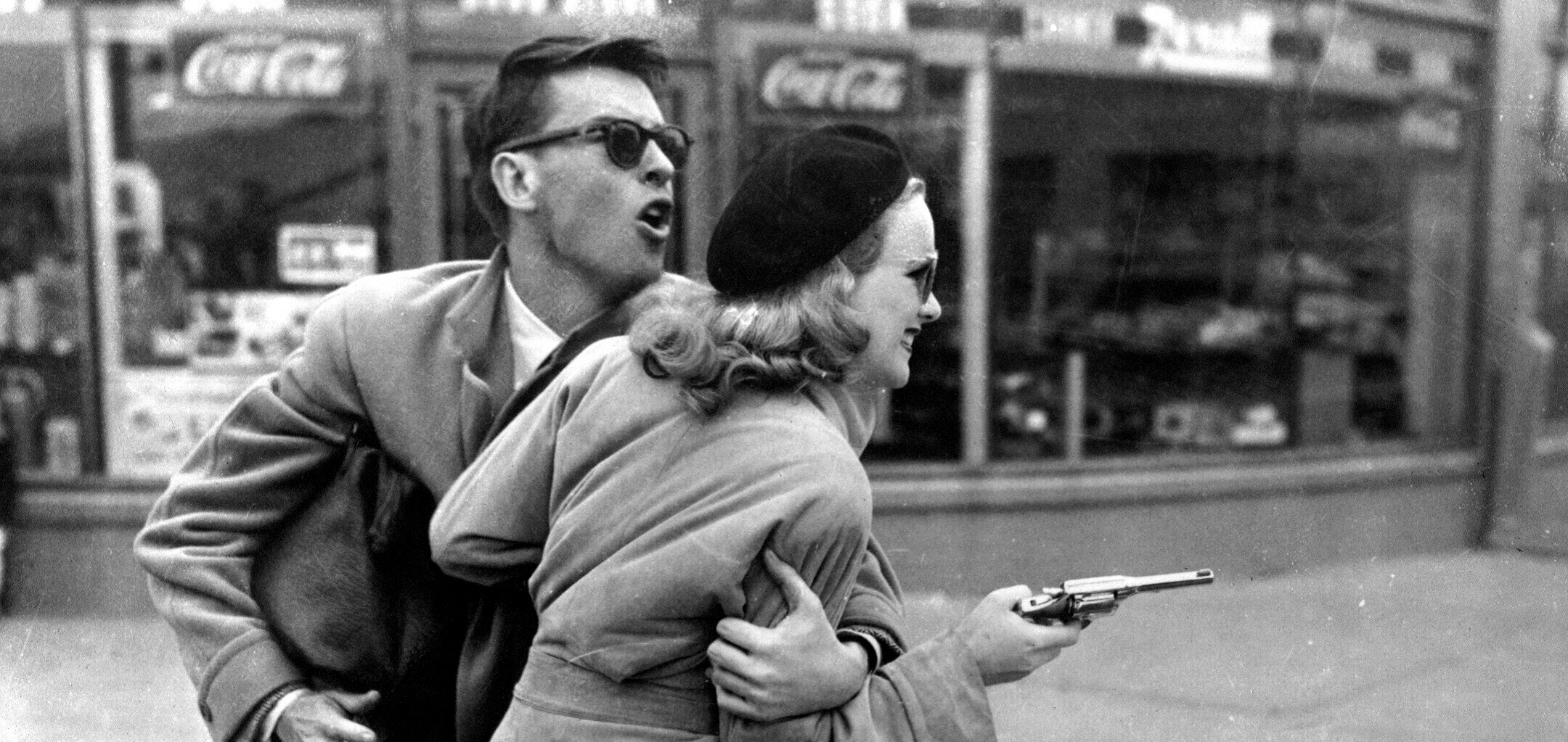 Peggy Cummins as Annie Laurie Starr and John Dall as Barton "Bart" Tare in a black and white still from Gun Crazy (1950). Annie is holding a revolver in the street while Bart is holding her back.