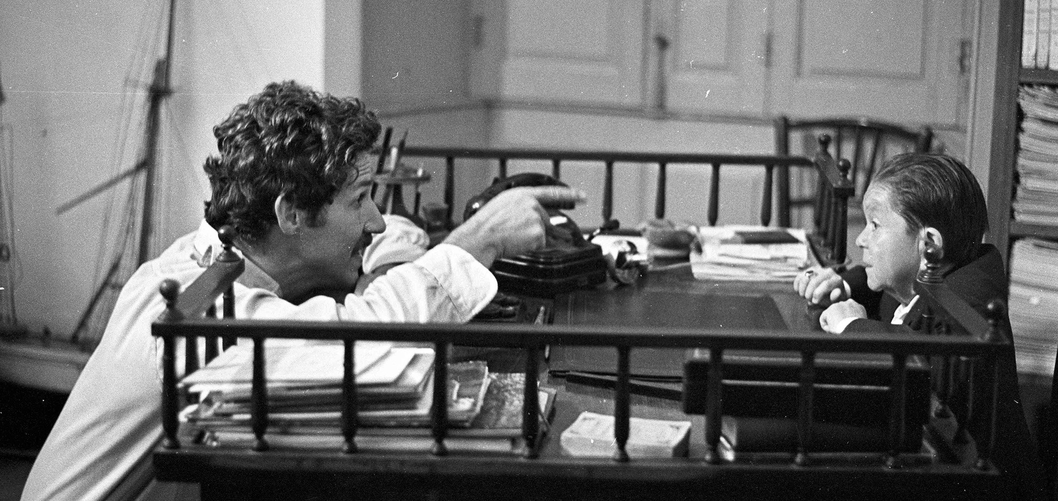 A black and white photo from the 1970s of director Werner Herzog sitting at a desk across from Helmut Döring (side view). Herzog is pointing at Döring