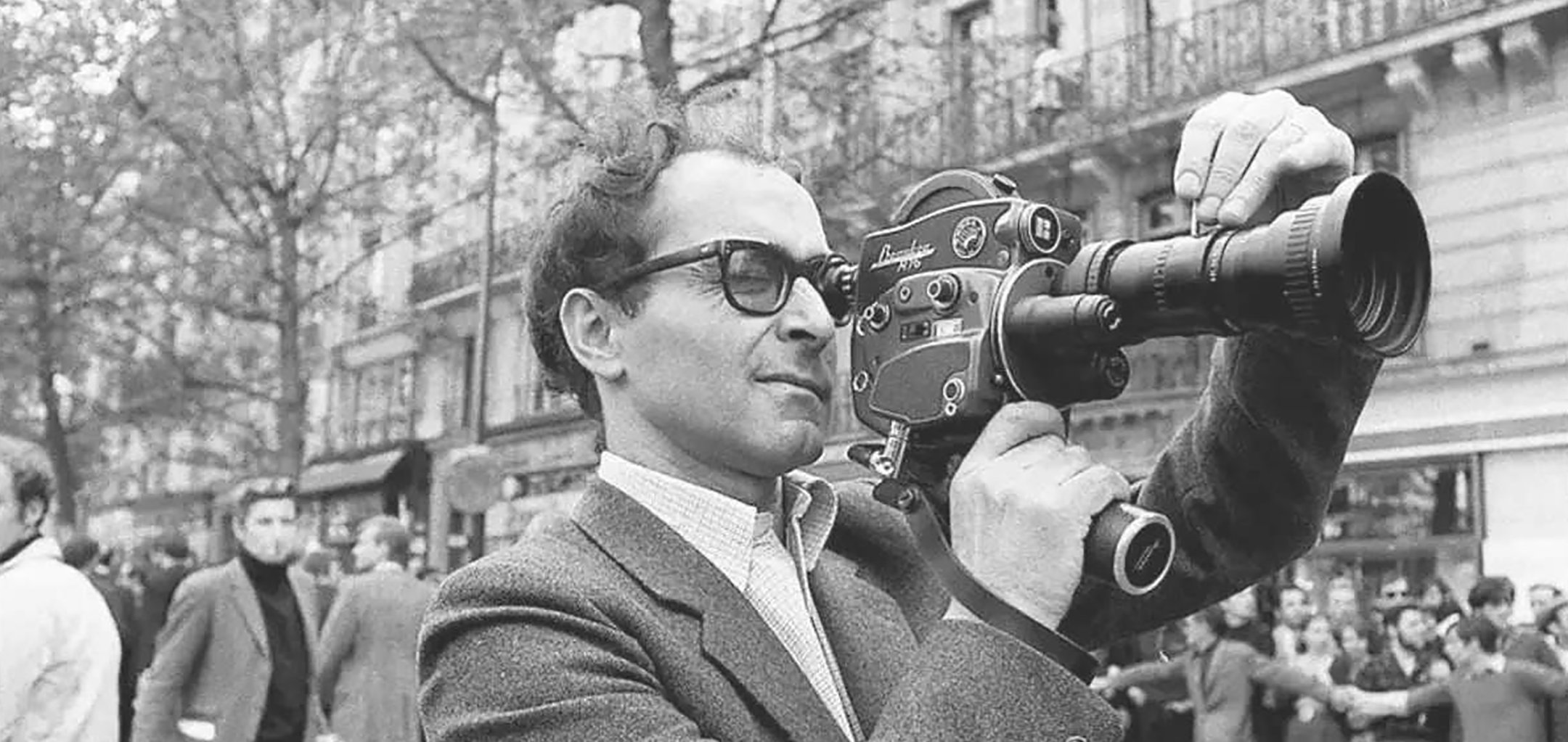 A black and white photo from the 1960s of a man (Jean-Luc Godard) in a suit and thick rimmed glasses holding a video camera on his shoulder.
