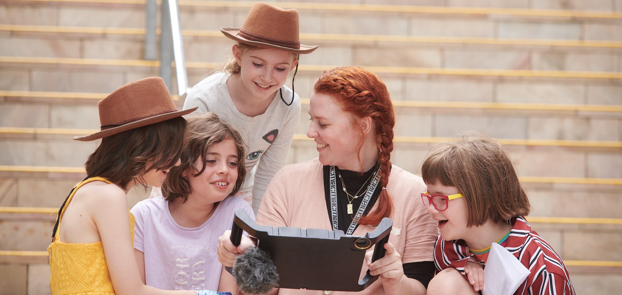 Four young girls look at a screen device with an ACMI facilitator. Two of the kids are wearing costume cowboy hats and they're all smiling. They are sitting on the stairwell at ACMI.