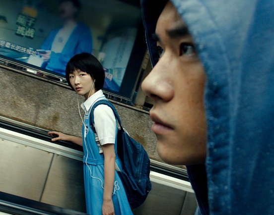 Jackson Ye in the foreground wearing a hood and Zhou Dongyu in the background on an escalator, in a still from 'Better Days' (2019) Courtesy of Magnum Pictures