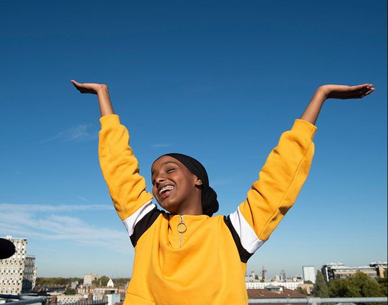 Kosar Ali as Sumaya in 'Rocks' (2017). Ali is wearing a black headscarf and yellow jumper. She is holding her arms up in joy, and her eyes are closed and she is smiling. It's a sunny day.