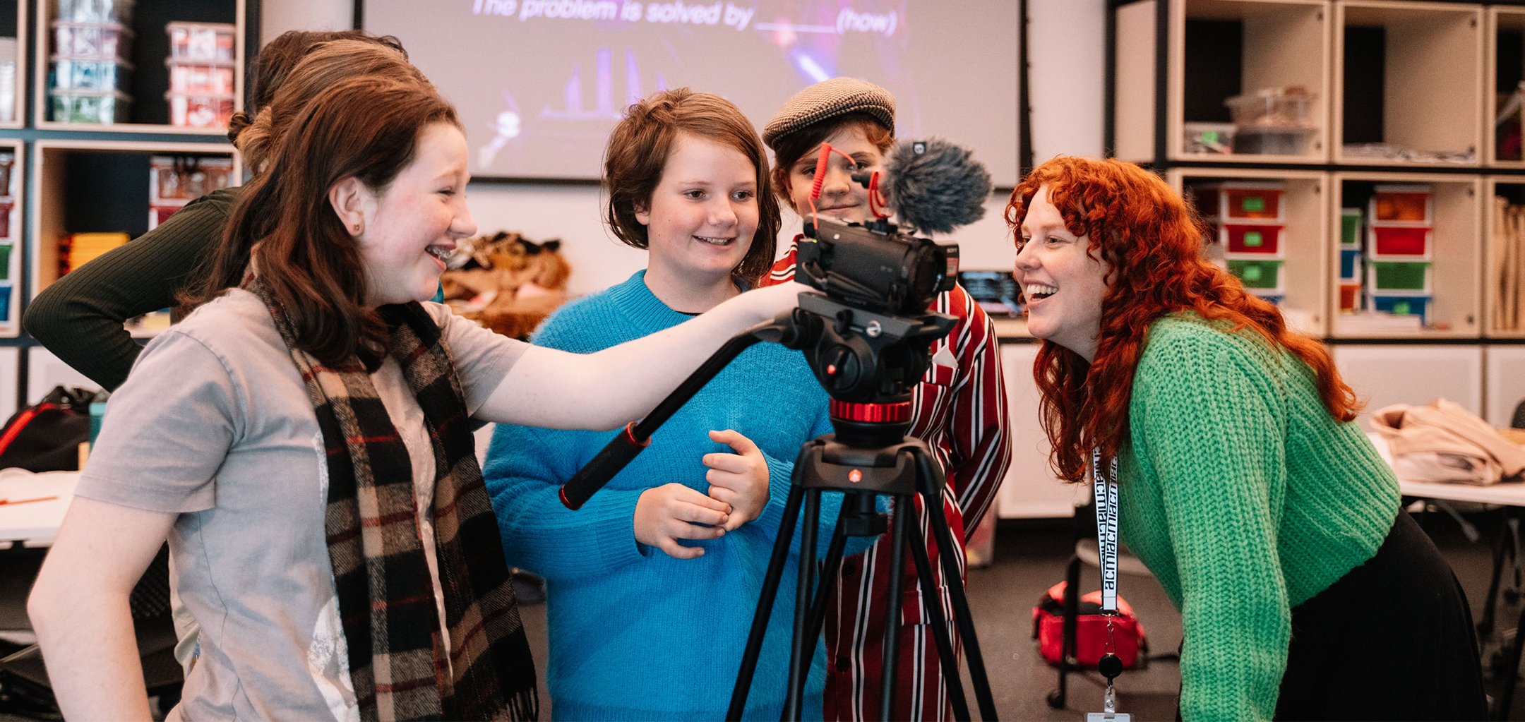 Four young teenaged girls smiling and operating a digital video camera on a tripod, with their ACMI facilitator