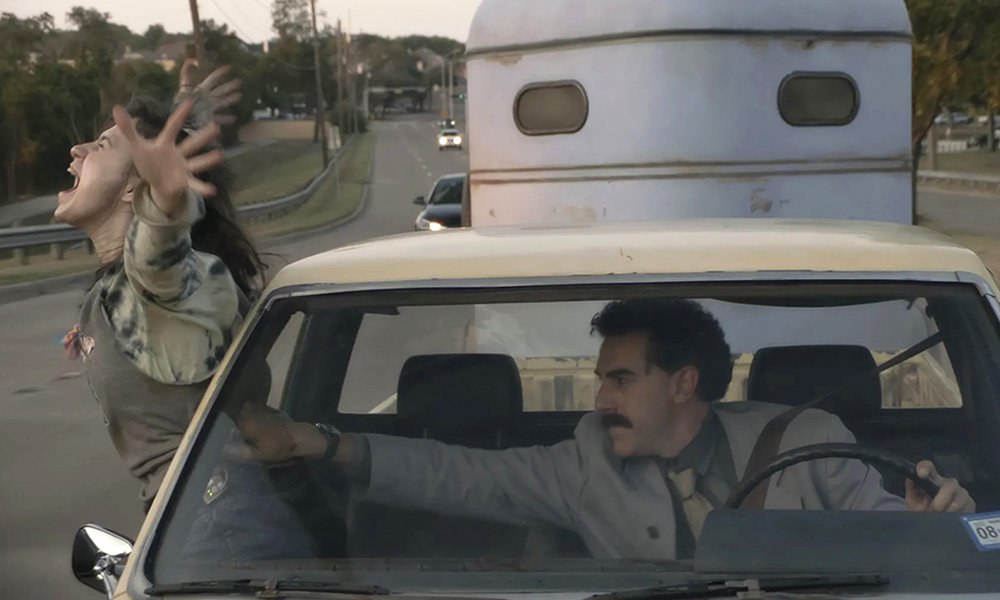 Maria Bakalova as Tutar leaning out a car window and Sasha Baron Cohen as Borat holding her back in Borat Subsequent Moviefilm (2020)