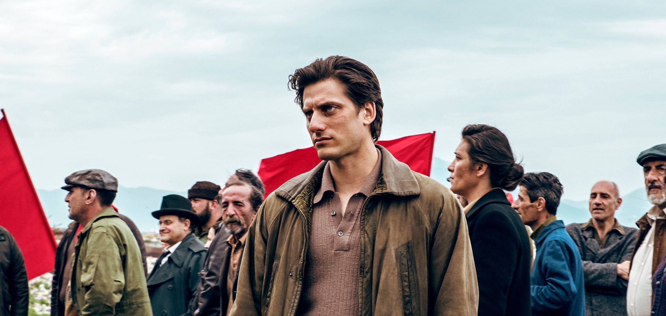 Luca Marinelli as titular character from Martin Eden, standing in front of a protesting crowd, some holding red flags