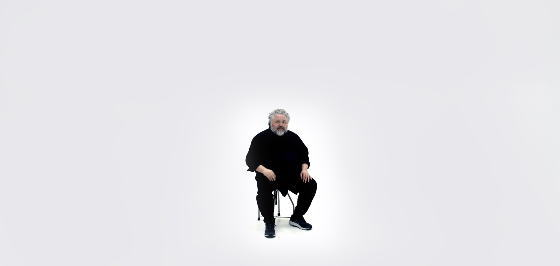 Designer Bruce Mau sits on a chair in a completely white room. He is framed centre and in the distance. He has short grey beard and short, curly grey hair, and he is wearing all black.