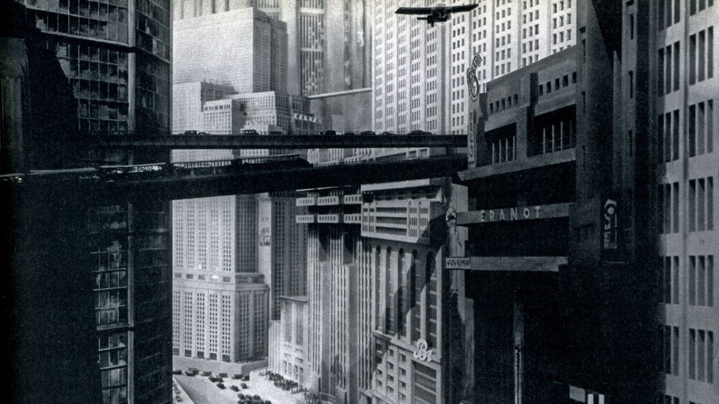 A dystopian cityscape in black and white, featuring looming buildings and bridges between them. A stream of cars snakes through the city below, while small planes fly through the sky.