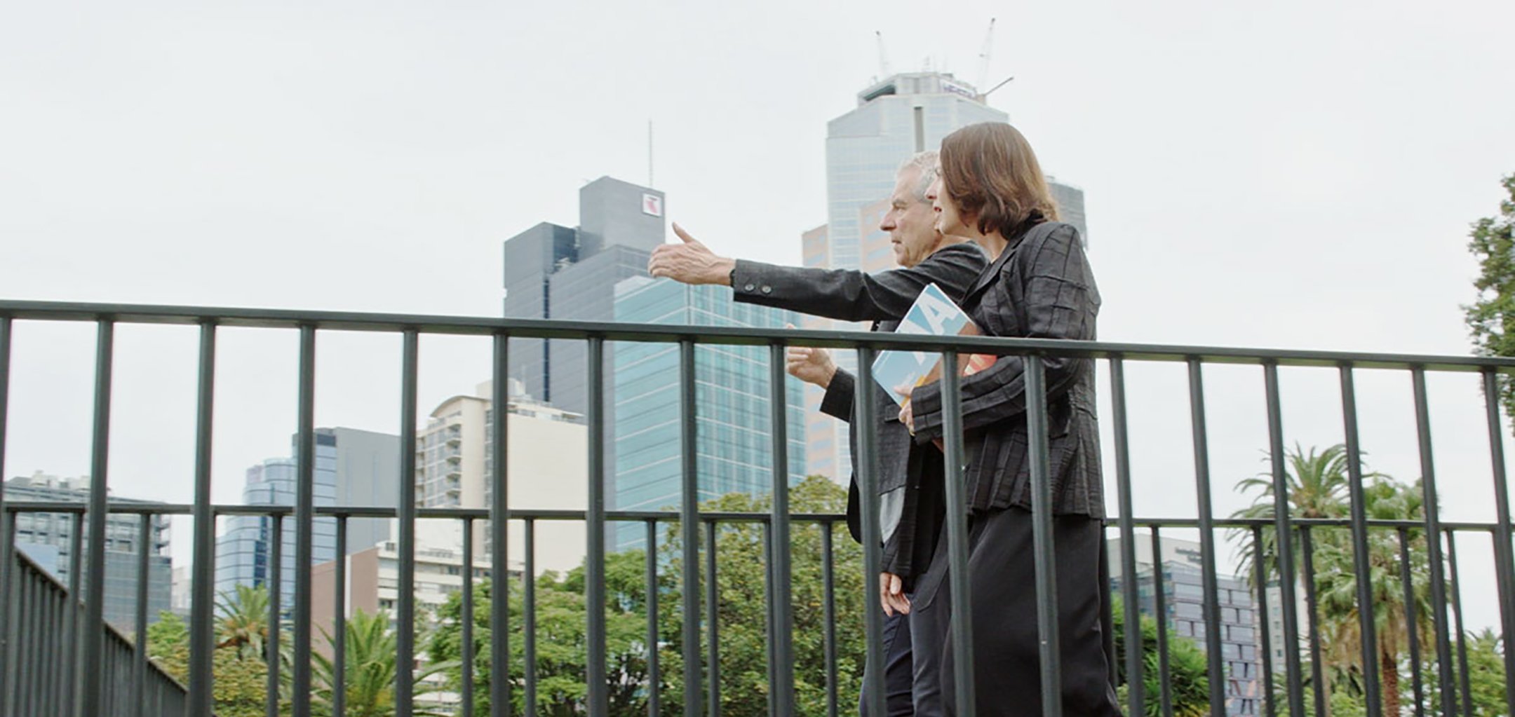 A man and a woman in corporate attire standing on a bridge in an inner city. The man is gesticulating and they are both looking off into the distance.