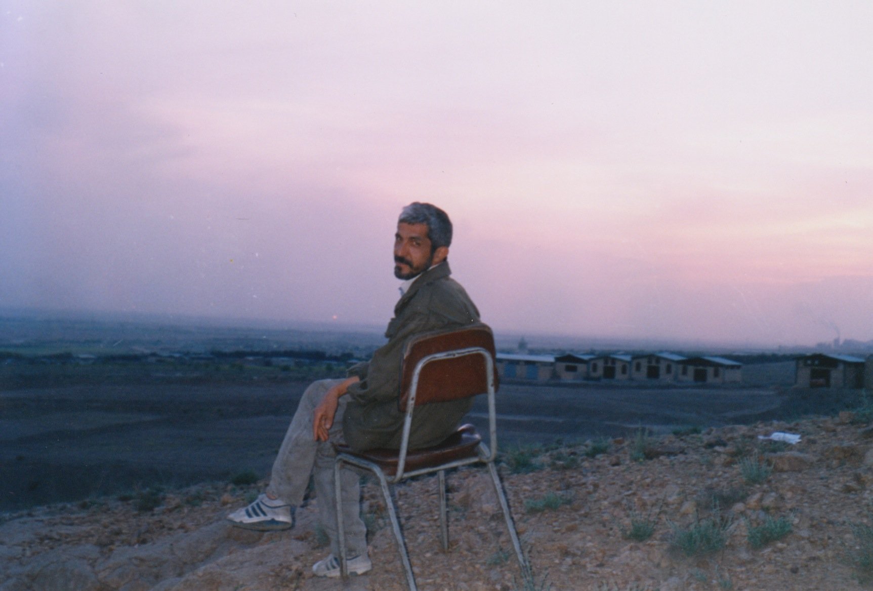 A middle-aged Iranian man sits in a chair overlooking a sparse landscape at dusk. He is looking back at the camera with a slight smile on his face.