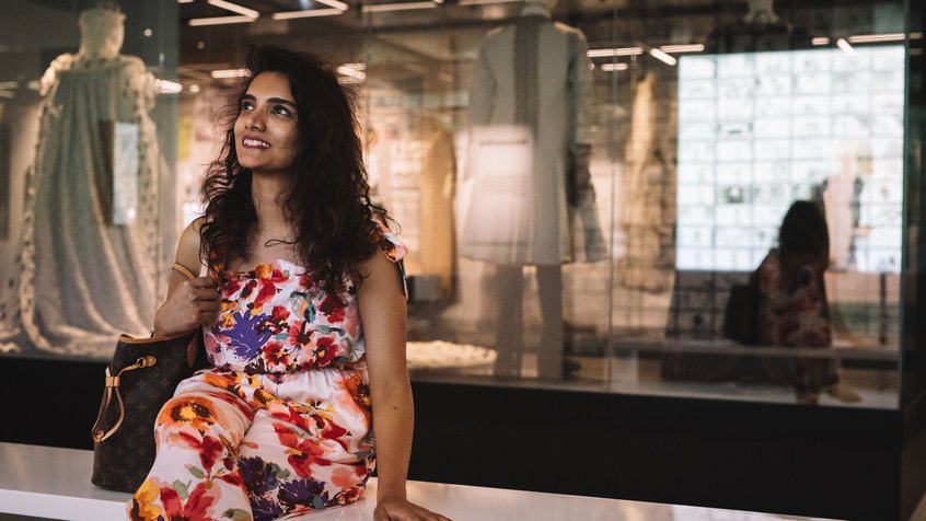A woman in a floral dress and long dark hair sitting on a bench and looking at an installation in The Story of the Moving Image (image credit: Phoebe Powell)