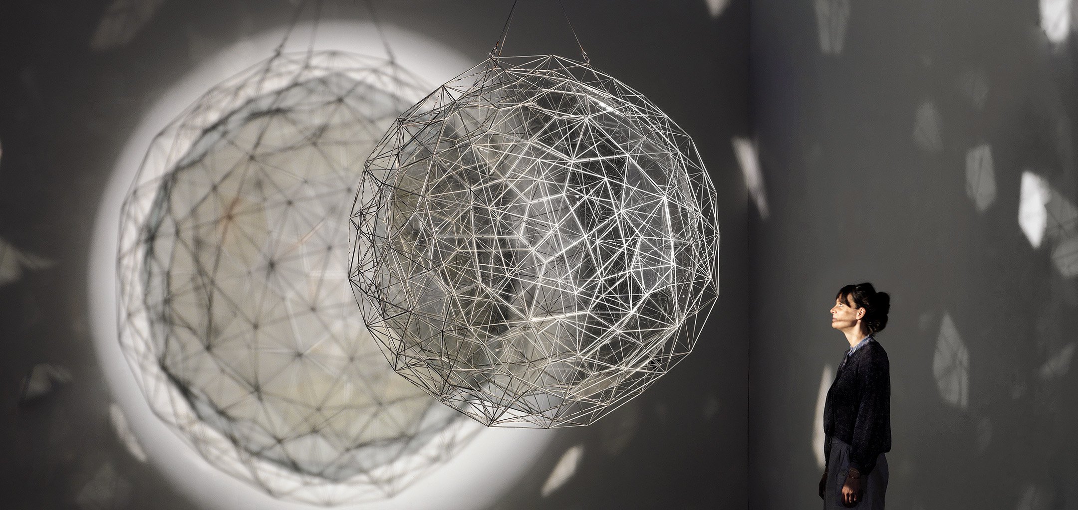 A woman looking at a large light-based art installation which is spherical and tessellated, and casting shadows and light on the gallery walls. Stardust particle, 2014, Olafur Eliasson, Tate: Presented by the artist in honour of Sir Nicholas Serota 2018 © Olafur Eliasson. Photo: Jens Ziehe