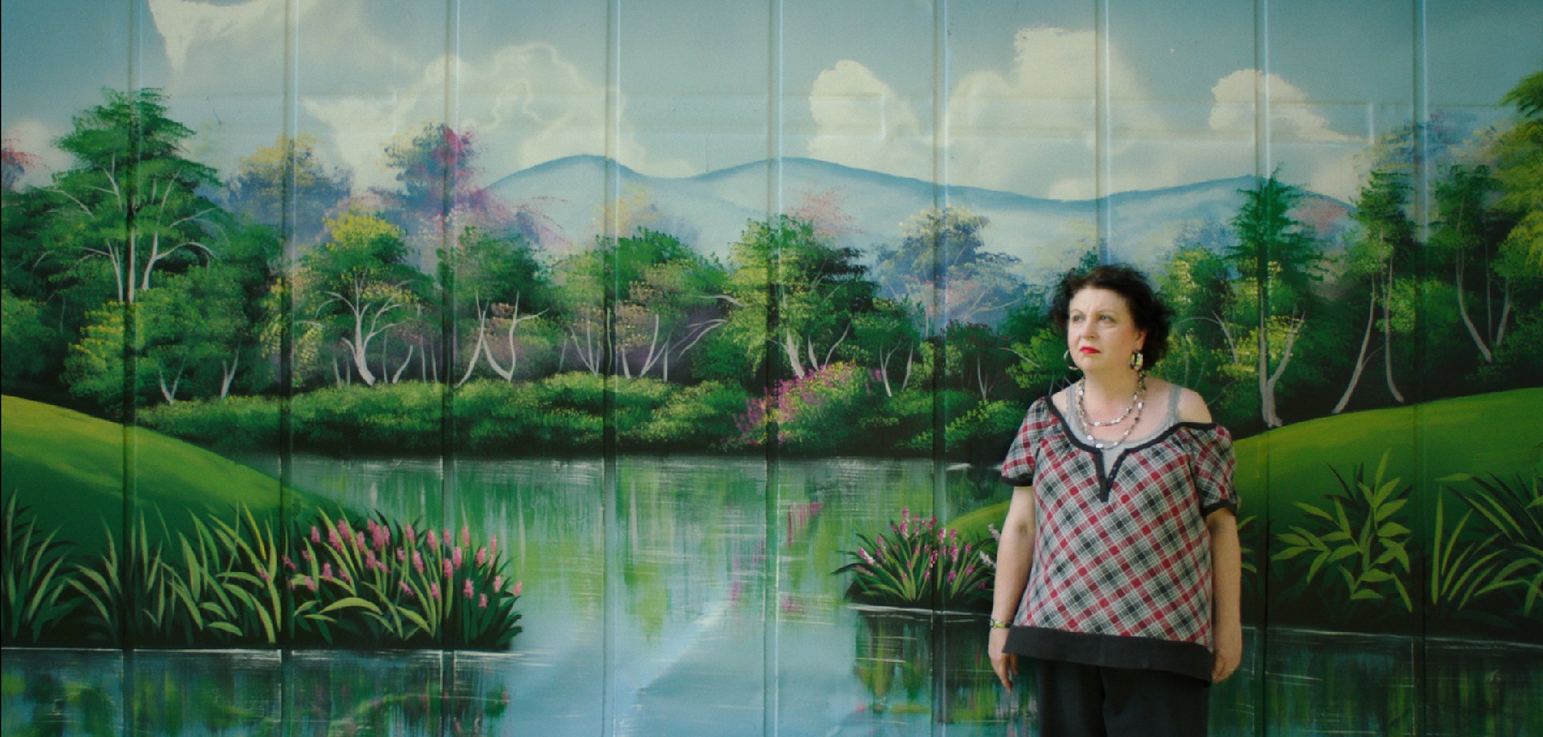 Barbara stands in front of a lake and garden setting painted on a wall in a still from 'Some Kind of Heaven' (2020)