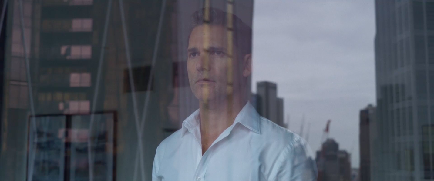 Eric Bana as Aaron Falk, staring out a window with a cityscape reflected on his face in a still from The Dry