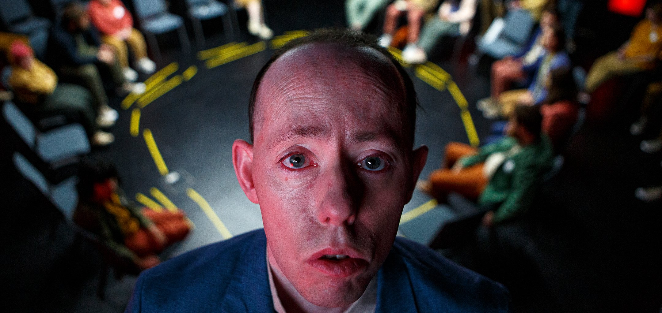 A person's solemn face is close to the camera, behind them is a circle of chairs with yellow taped marks on the floor. Some chairs are occupied by people.