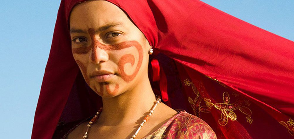 Closeup of a woman in a red headscarf and red and gold Colombian Indigenous dress. Still from Birds of Passage (2018)