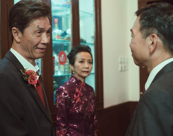 Tai Bo and Ben Yeun meet each other at an event, with Tai Bo's wife in the background, still from 'Suk Suk' (2019)